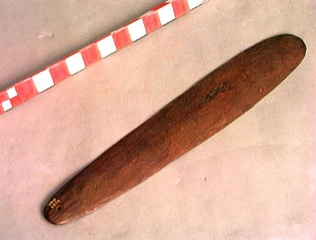 A long red-brown bull-roarer blade incised from end to end with overlapping concentric-circle motifs, a single small perforation drilled near...