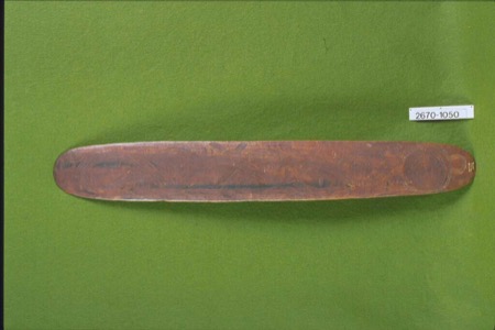 A broad board, photographed on green felt, its face incised with a continuous interlocking wave pattern — an Aboriginal Australian bull-roarer...