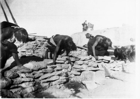 Five Hopi priests kneel doubled over along the stone edge of a kiva roof, heads down and hair hanging, vomiting after taking the emetic.