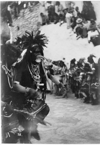 A Hopi snake priest in a feathered headdress and shell necklaces holds a large snake crosswise in his mouth, its body looped past his cheek; a second priest steadies him at the shoulder while spectators watch from the rooftops behind.