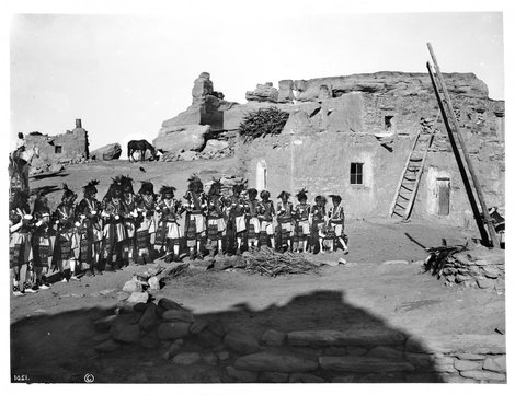 Seventeen Hopi Antelope priests in patterned kilts and feathered headdresses file out of a stone kiva onto open ground, a mounted man watching from behind at left.