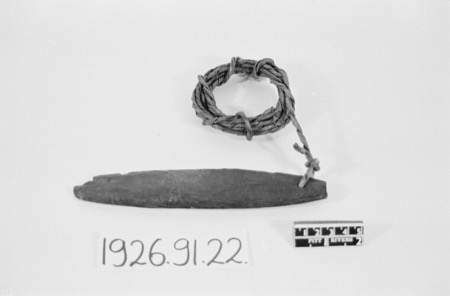 A small, plain dark blade with a coiled cord looped through a hole at one end, photographed on a museum scale card: an African bullroarer of...