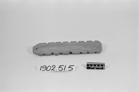 A notched wooden board with a leaf-shaped incised spine and a hole at one end, photographed against a museum scale card: an English bullroarer...