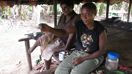 Nakre Abia sits with an arm extended as she swings a coconut-leaf frfr; another person sits behind her on the raised platform.