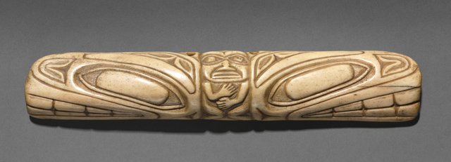 Carved bone tube, a Tlingit shaman's charm: a human face at the center gripped between two animal heads, with formline ovoids and rib-like panels along the body.