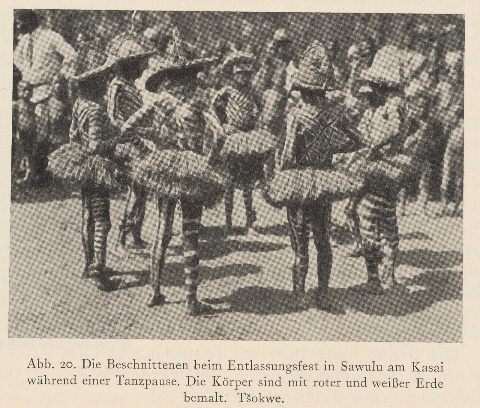 Chokwe initiates with striped and geometric body paint stand in a circle wearing grass skirts and crested hats.