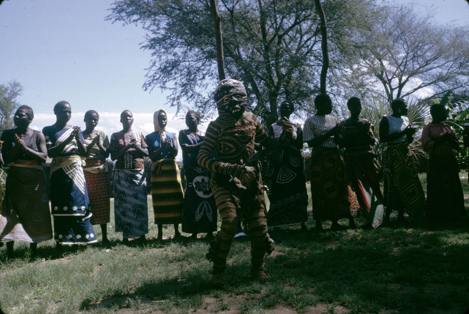 Luvale Makishi dancer in a patterned full-body costume performs before a line of clapping women in Lusaka.