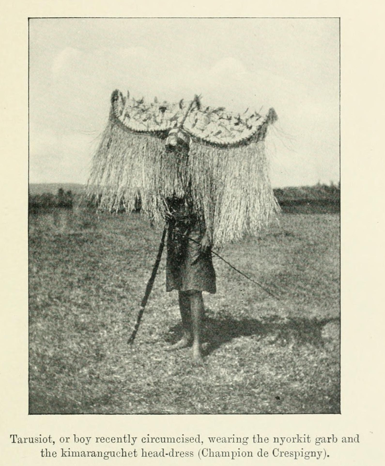 Nandi initiate in nyorkit clothing wears a broad crescent-shaped headdress fringed with long hanging fibres.