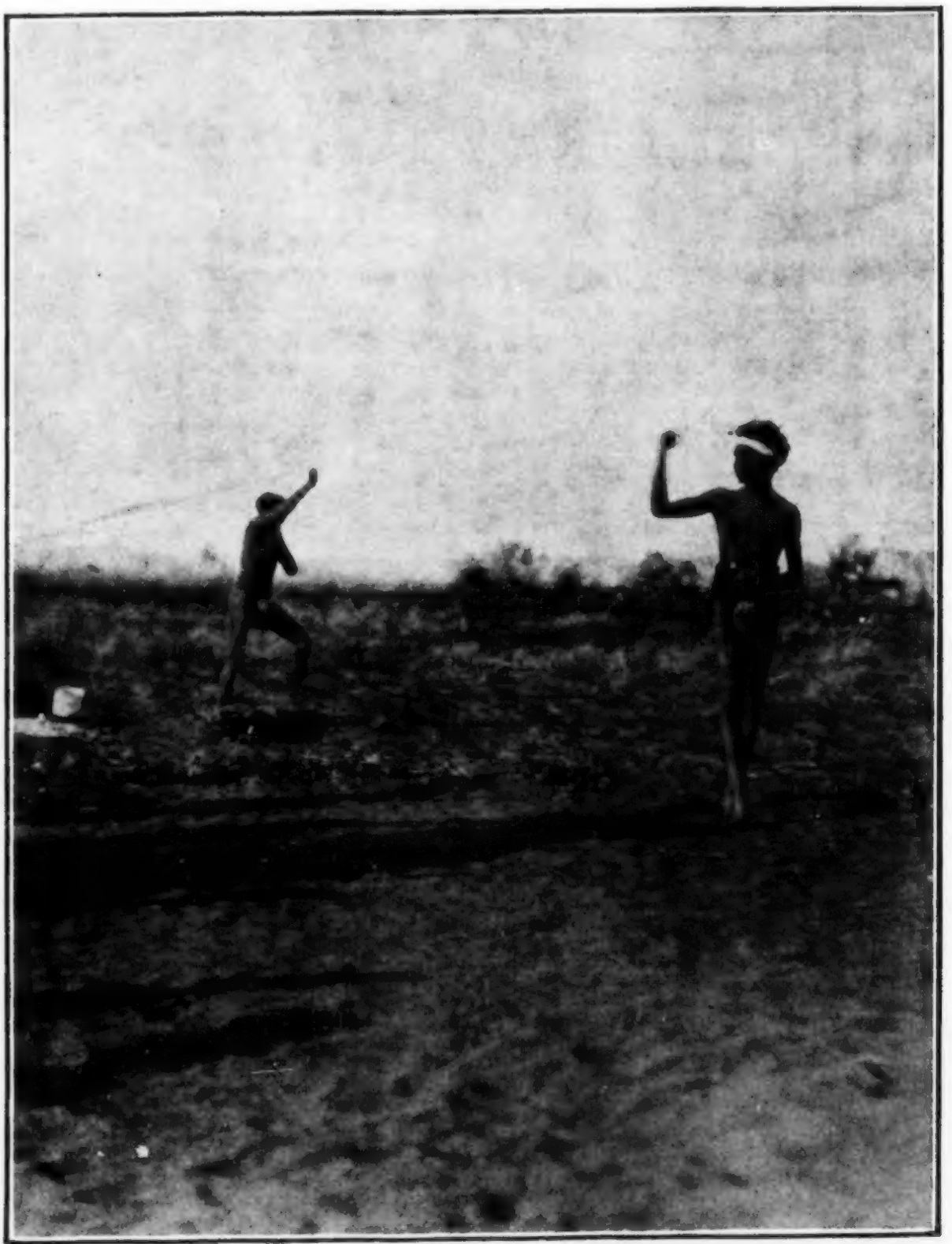 Two Karajarri men stand apart on open ground with raised arms as they swing bullroarers.