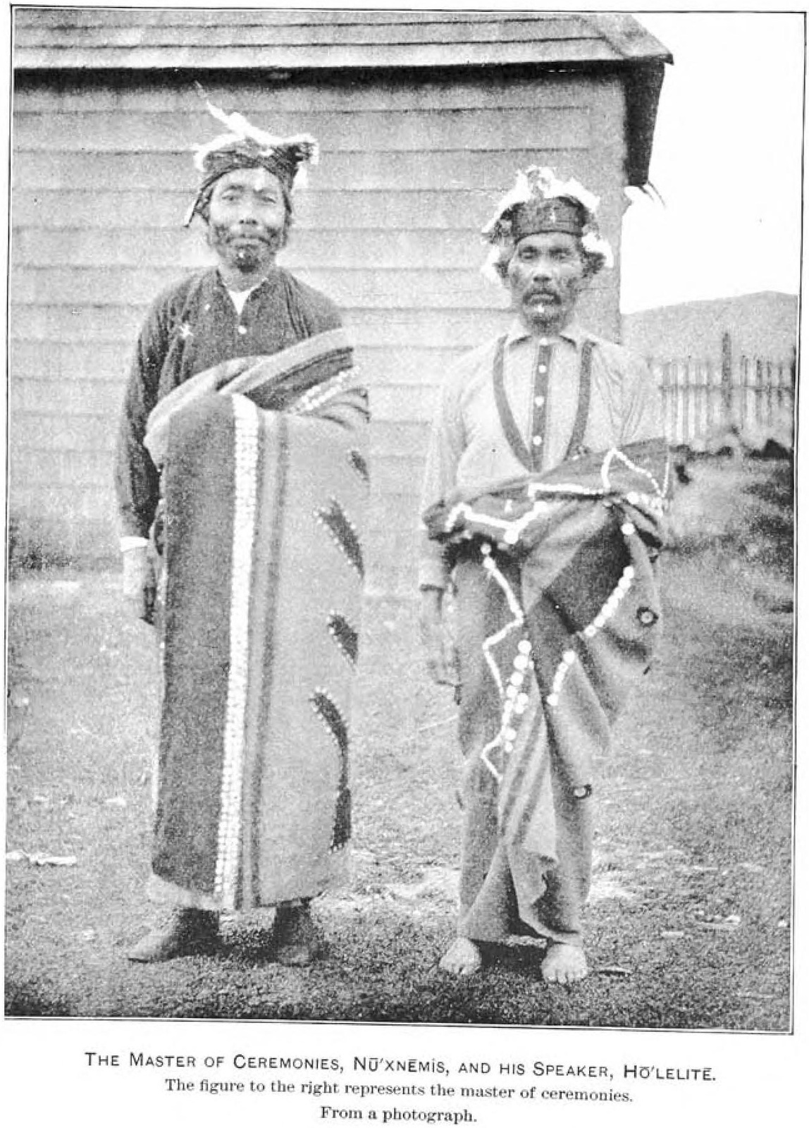 Kwakwaka’wakw Winter Ceremonial master Nu’xnemis and speaker Ho’LElite stand in feathered headrings and patterned blankets.