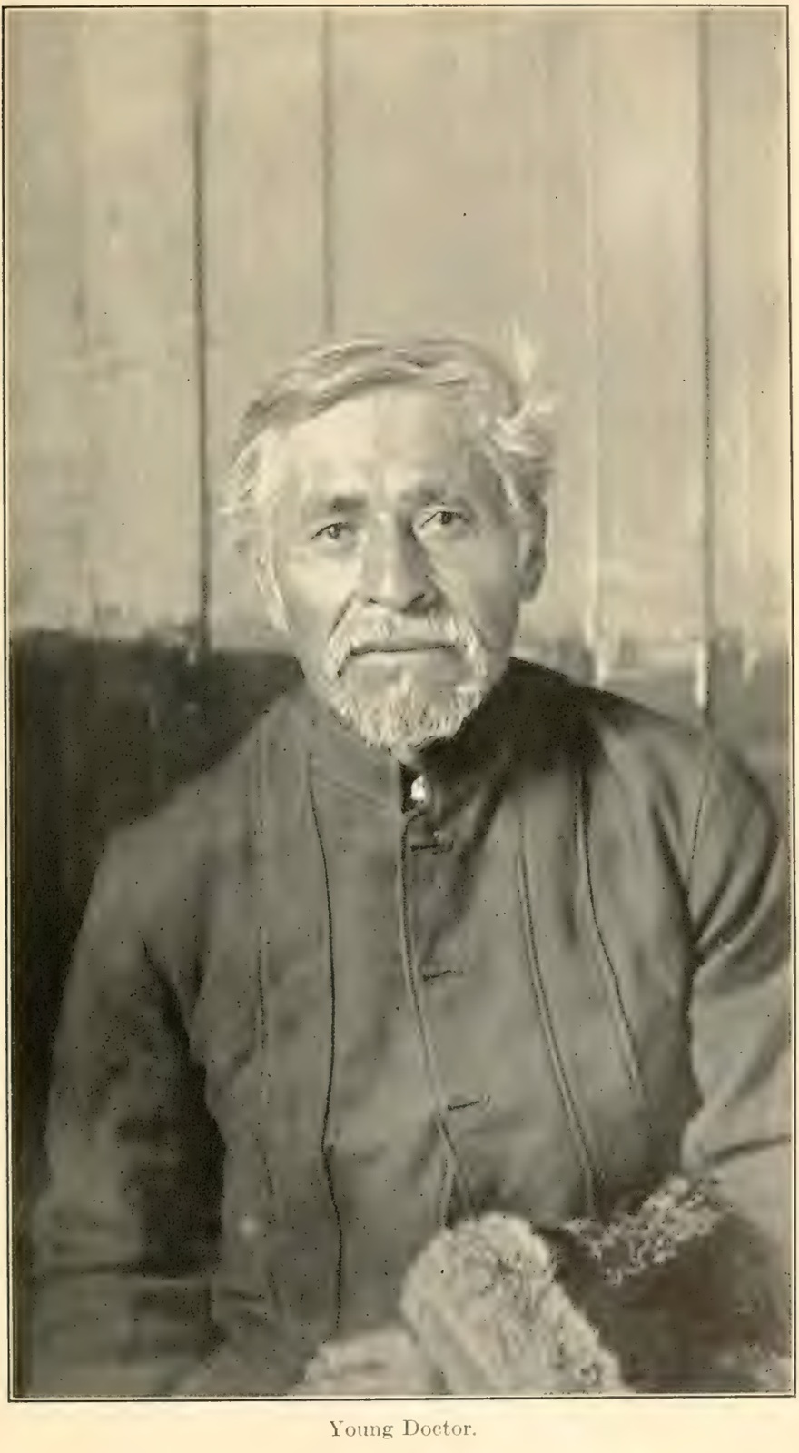 Makah elder Young Doctor sits before a plank wall, wearing a dark buttoned jacket, with grey hair, moustache and beard.