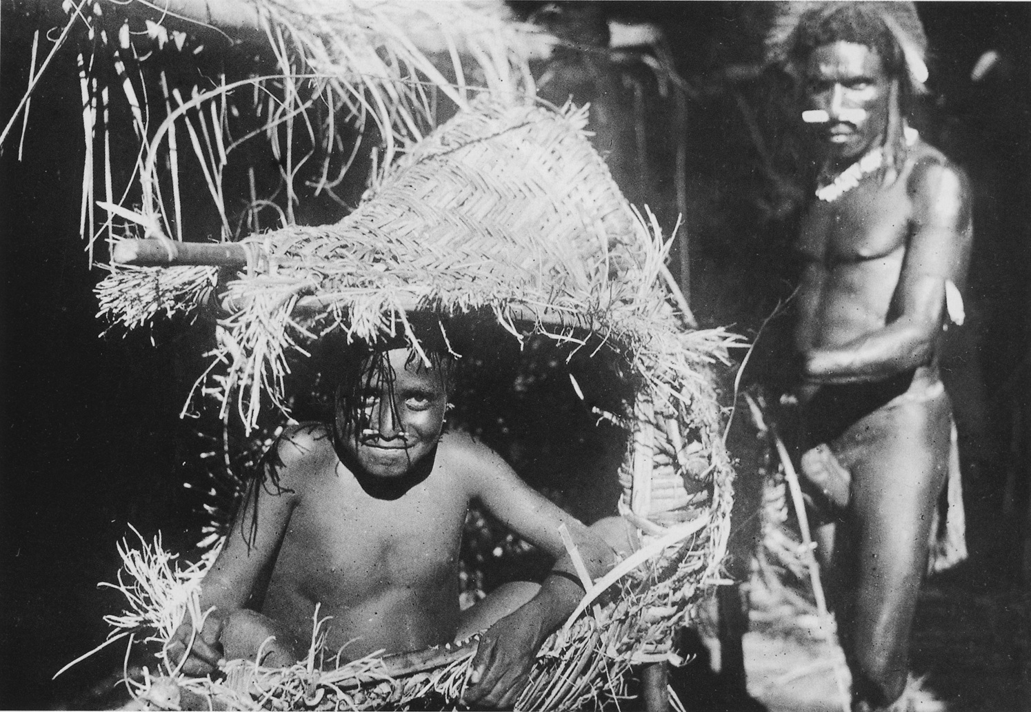 Ove Eh sits within the open woven mouth of a kaiaimunu, with an adult working beside him.
